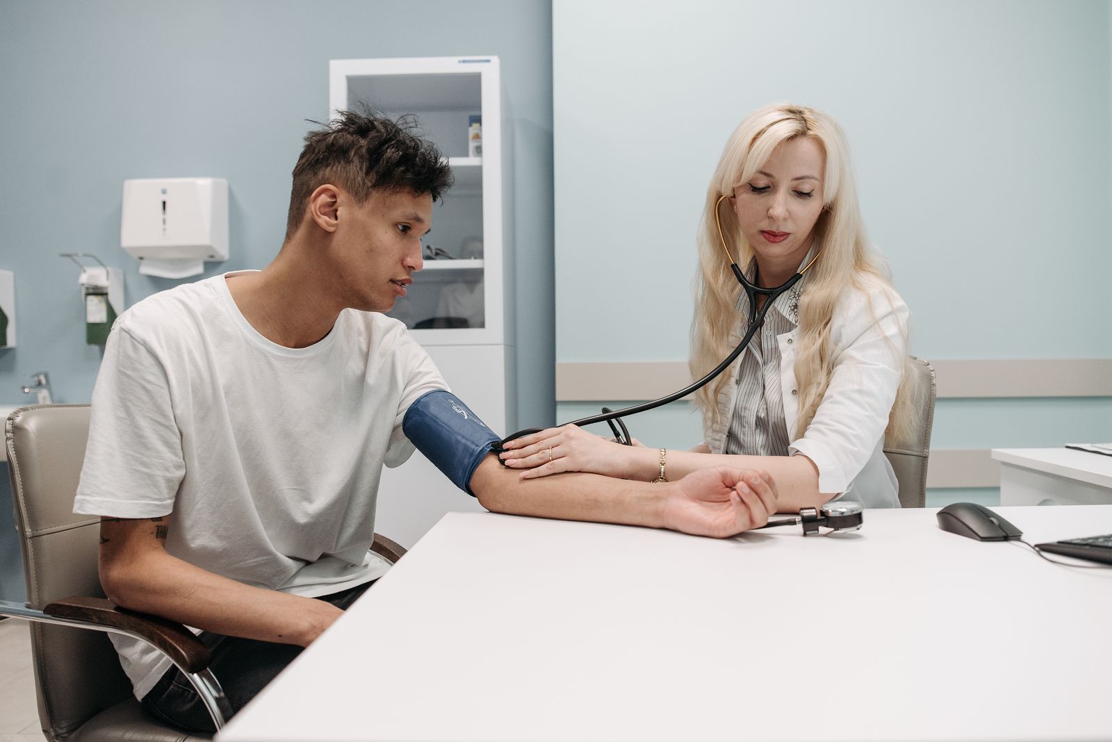 A clinician taking a patient's blood pressure with a cuff and stethoscope at a clinic desk.
