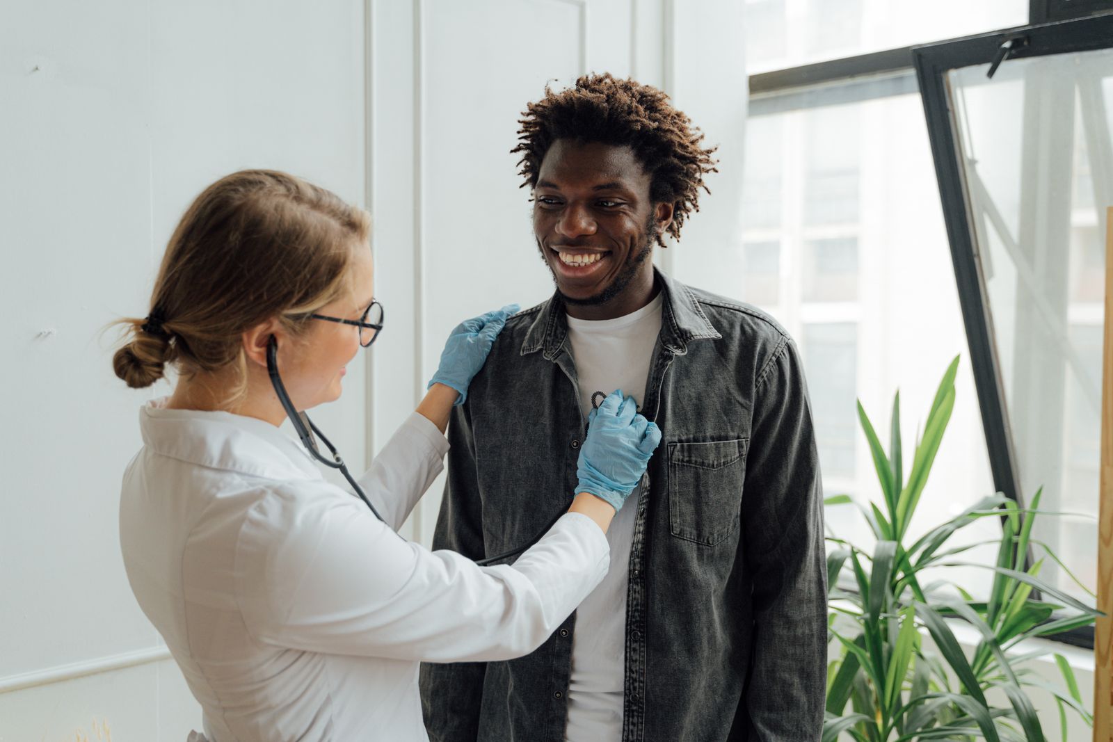 A clinician in a white coat and gloves listening to a smiling man's chest with a stethoscope.